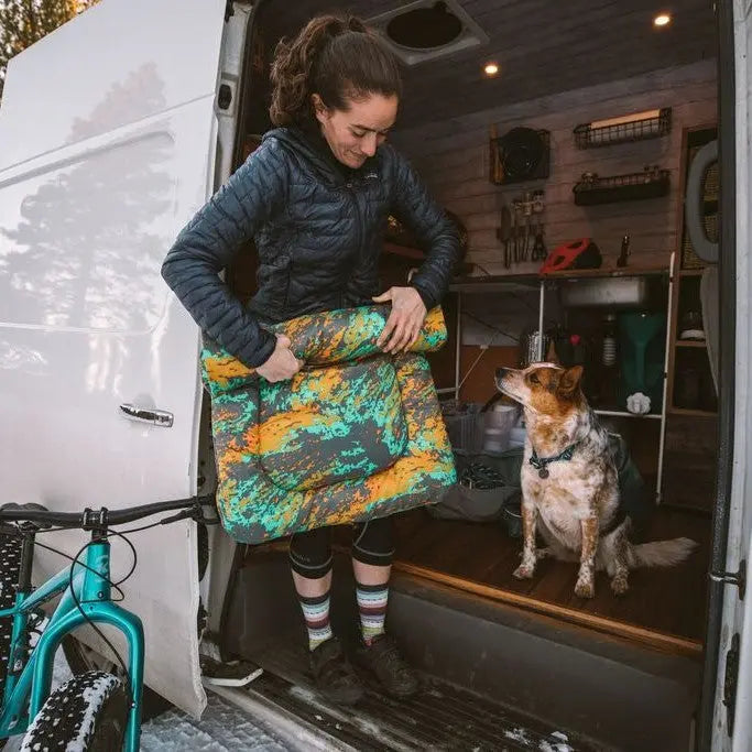 Woman carrying a colorful dog bed from a van, with a curious dog watching nearby.