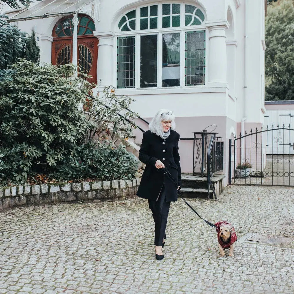 Woman in a black coat walking a dog on a leash outside a stylish home.