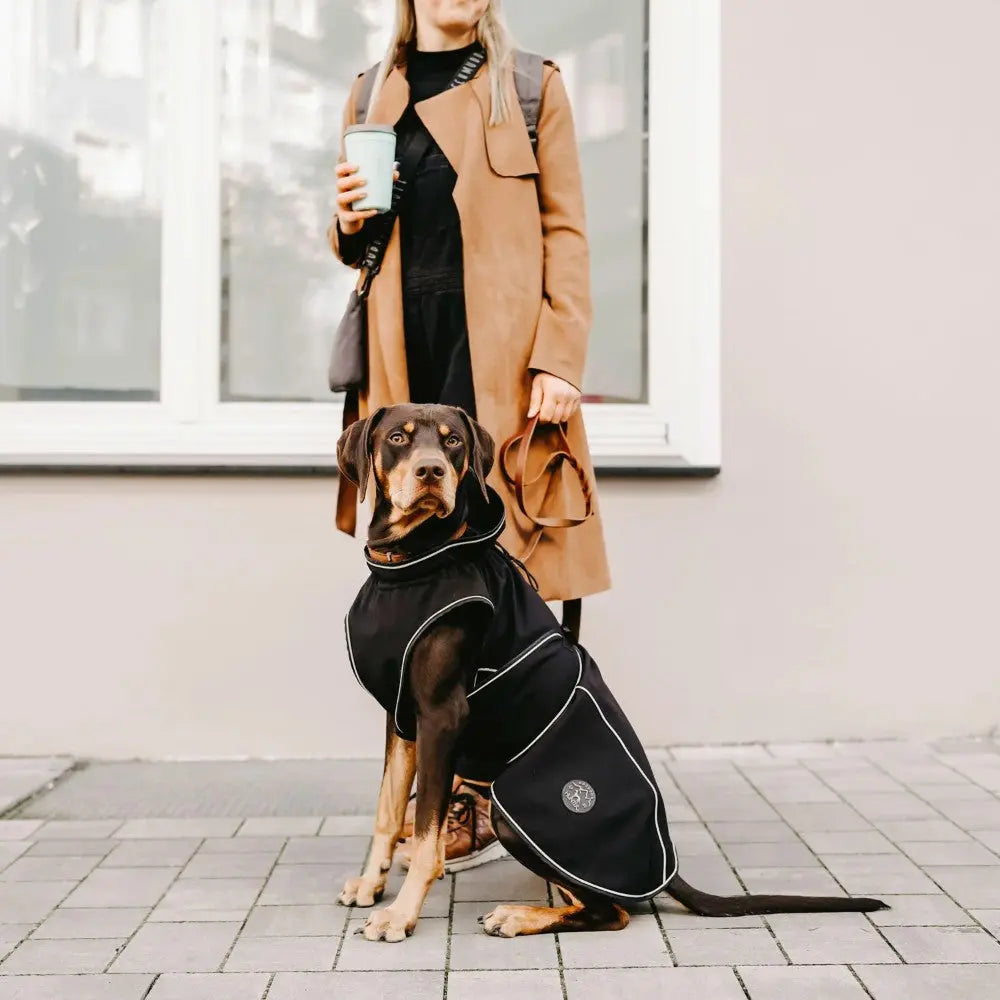 Stylish dog in a black coat sits beside a woman holding a coffee cup on a city sidewalk.