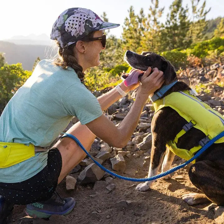 Hiker in a cap and sunglasses lovingly interacts with a dog in a bright yellow life jacket on a rocky trail.