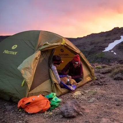 Couple enjoying a cozy evening in a Marmot tent with a dog at sunset in a scenic outdoor setting.
